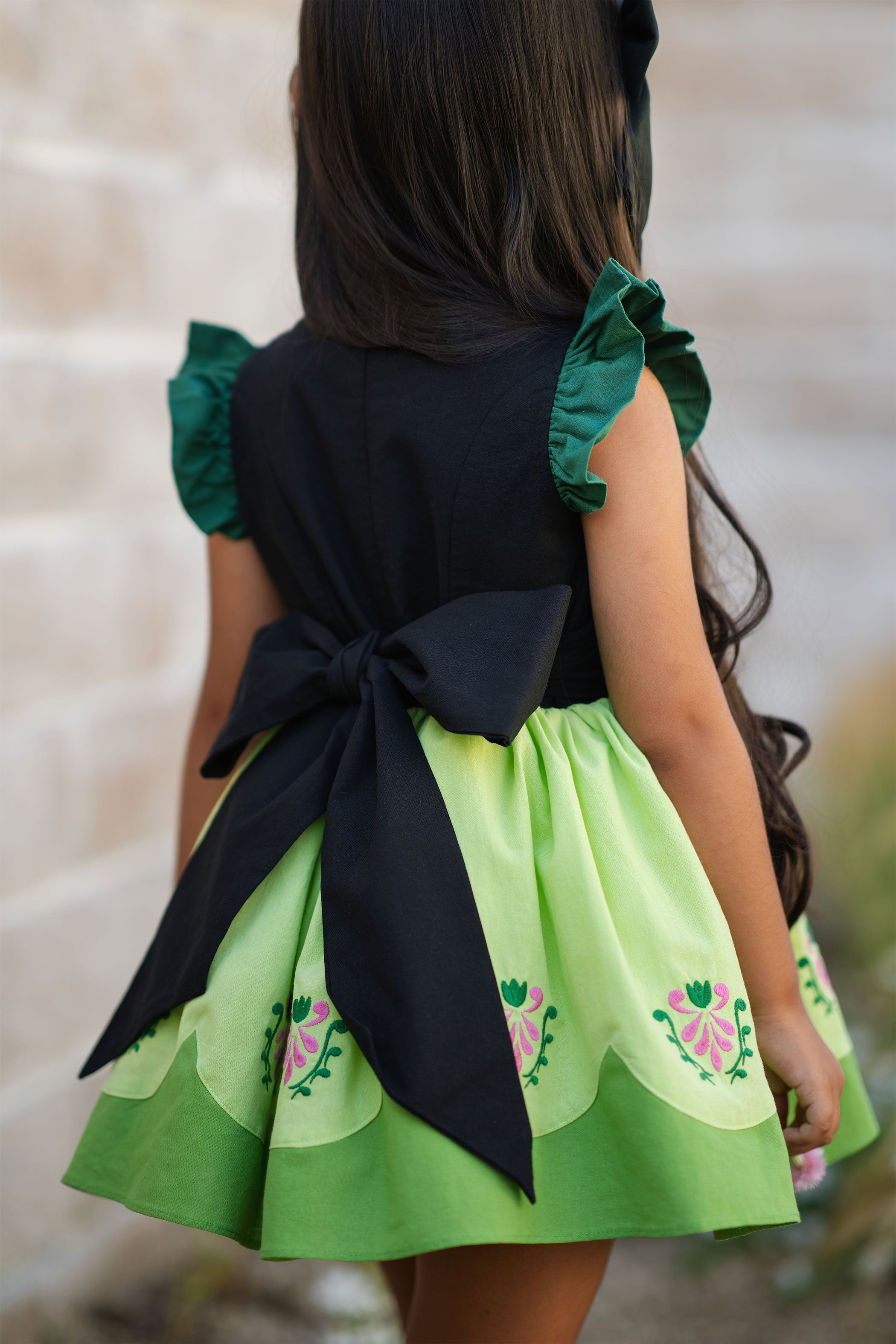 Green dress with floral patterns and a black bow on the back, worn by a child.