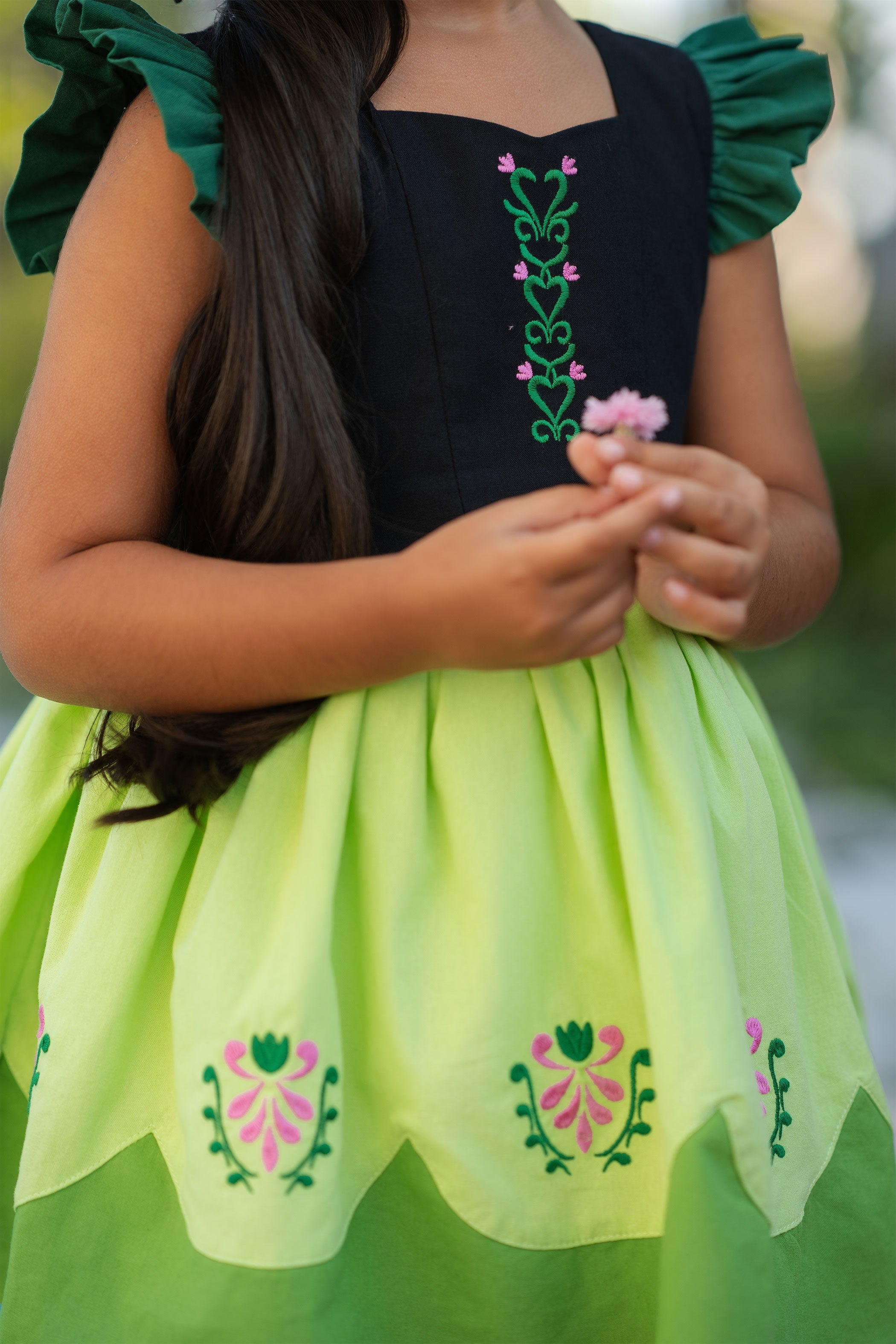 Child wearing a black top and green skirt with floral embroidery, holding flowers.