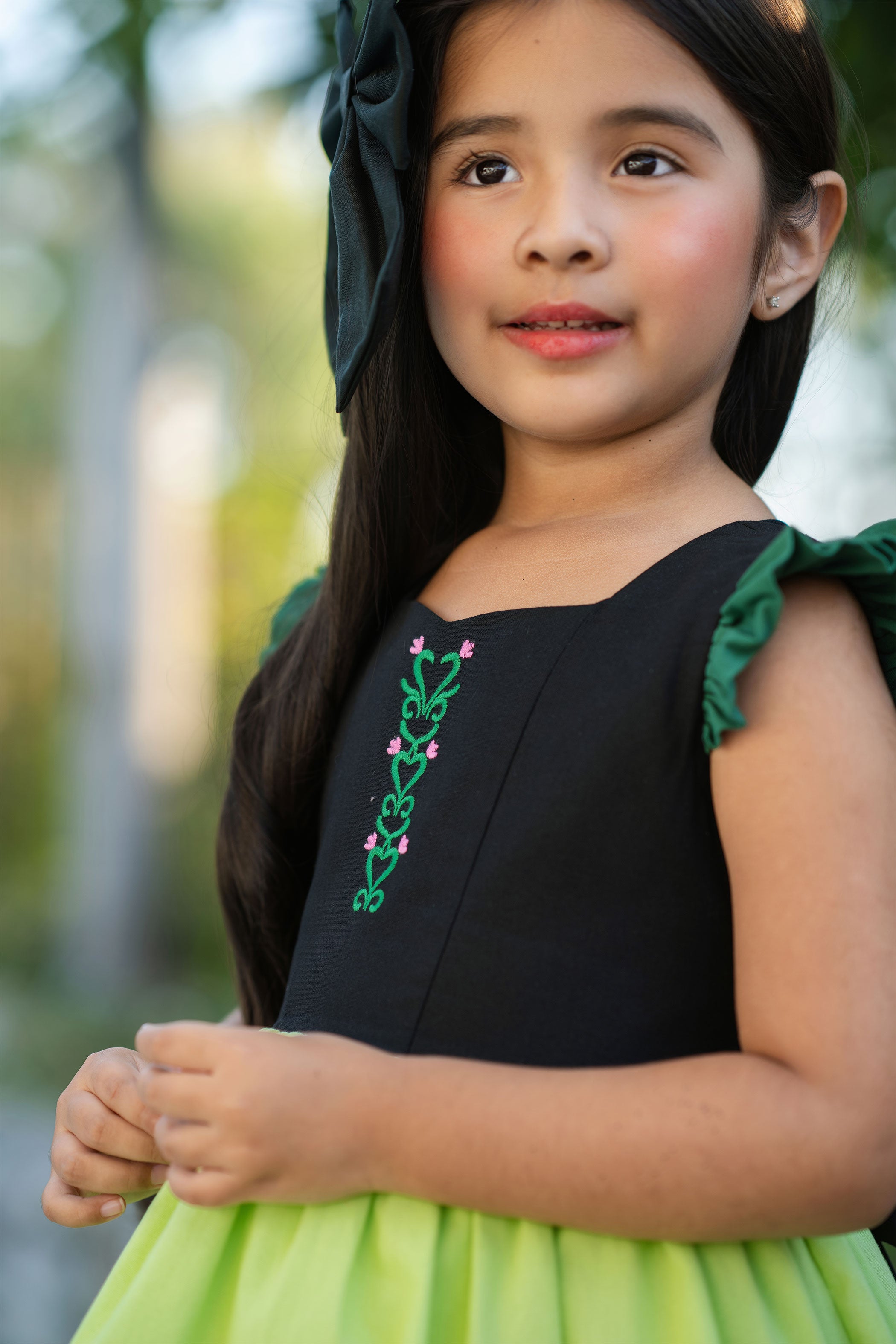 Young girl wearing a black and green dress with floral embroidery outdoors.