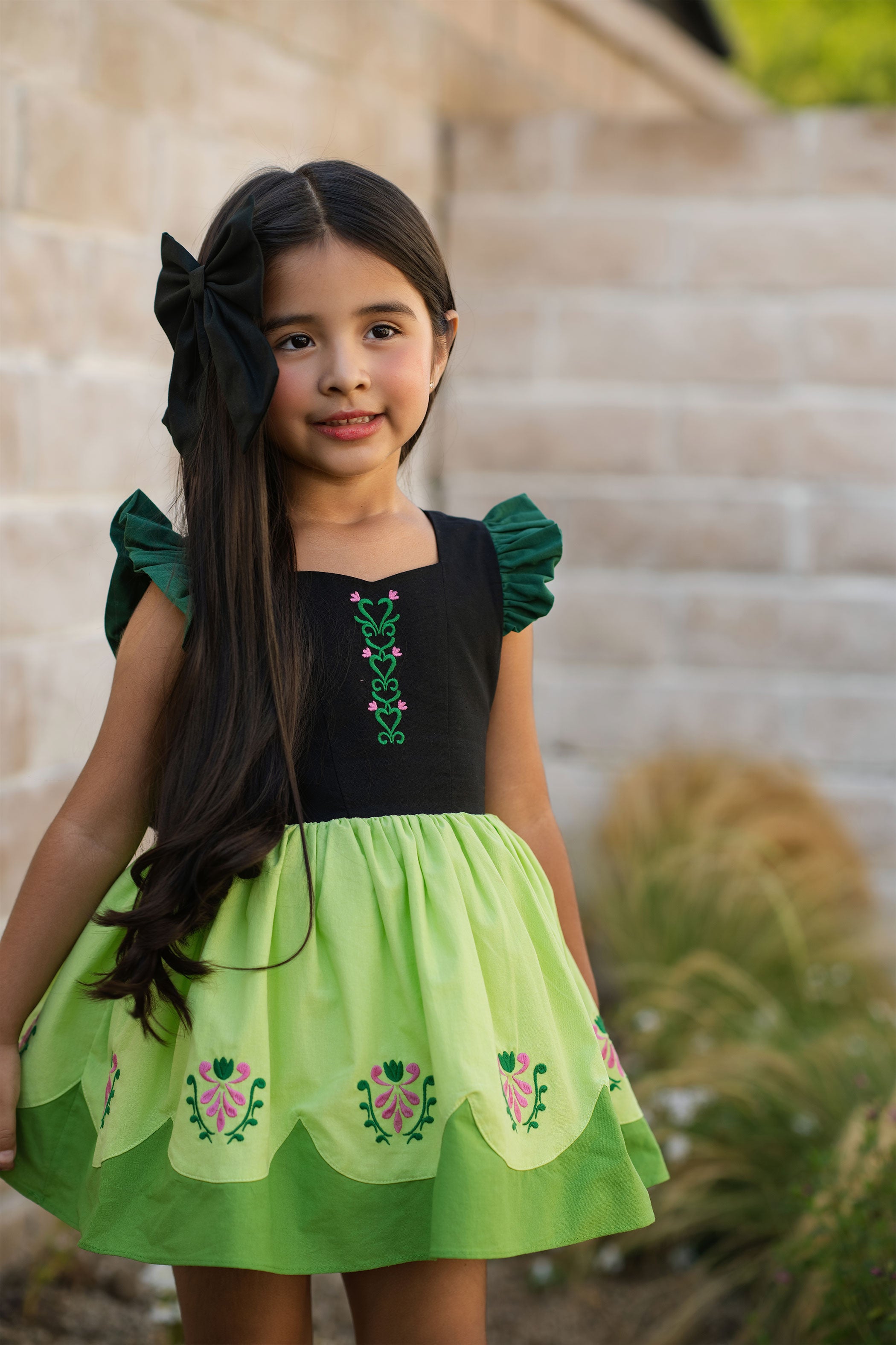 Young girl wearing a green and black dress with floral embroidery, standing outdoors.