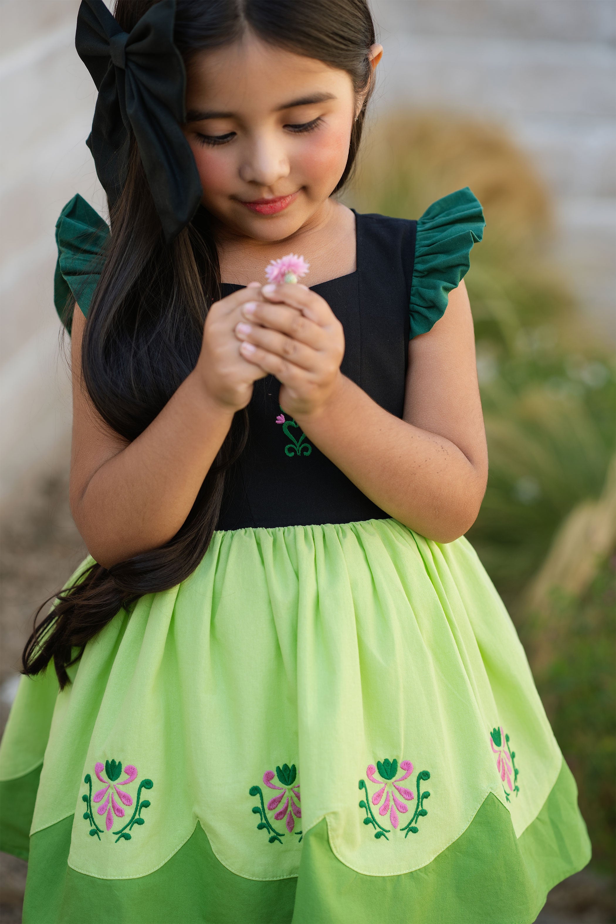 Young girl in a green and black dress holding a flower outdoors.