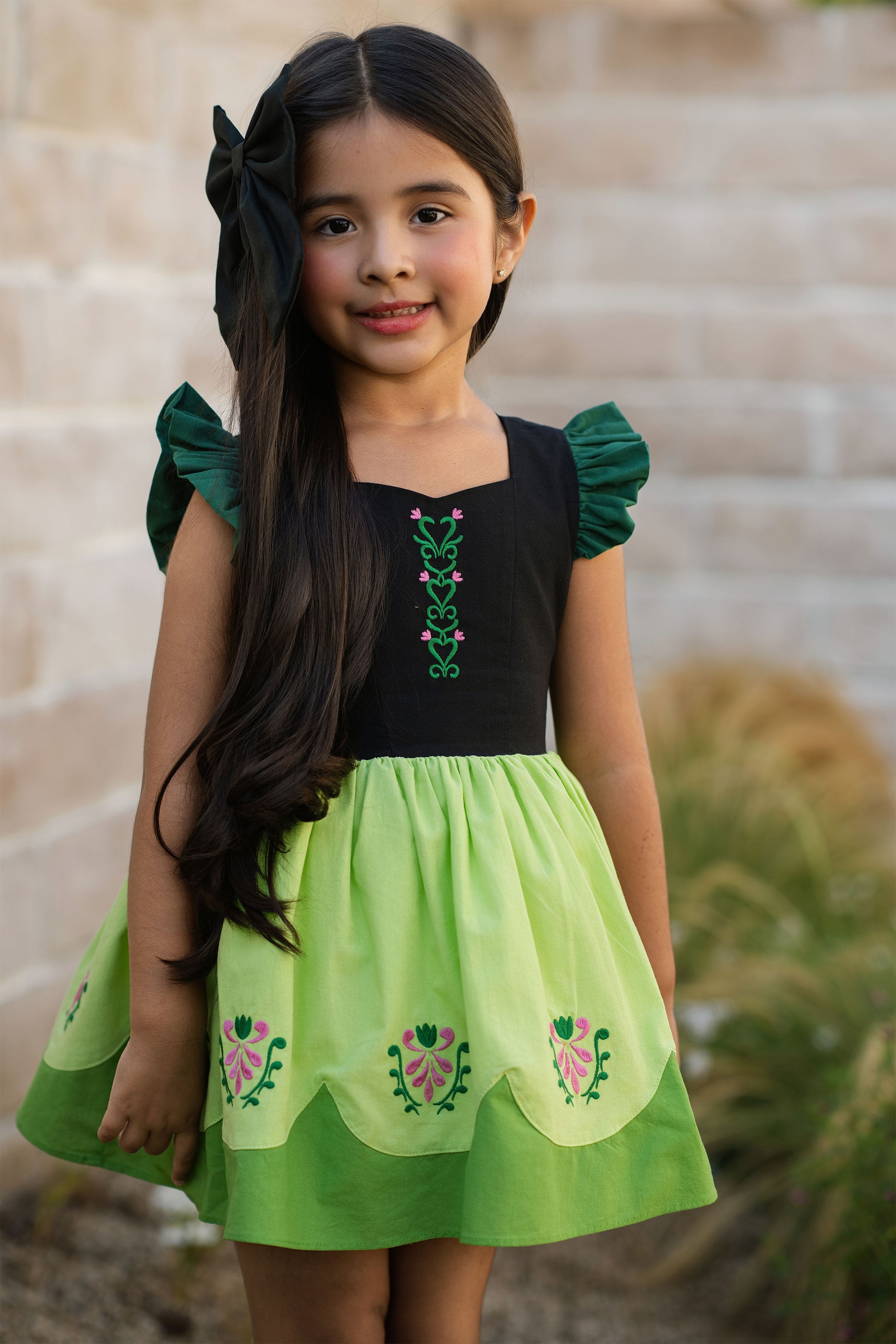 Young girl wearing a green and black dress with floral embroidery, standing outdoors.