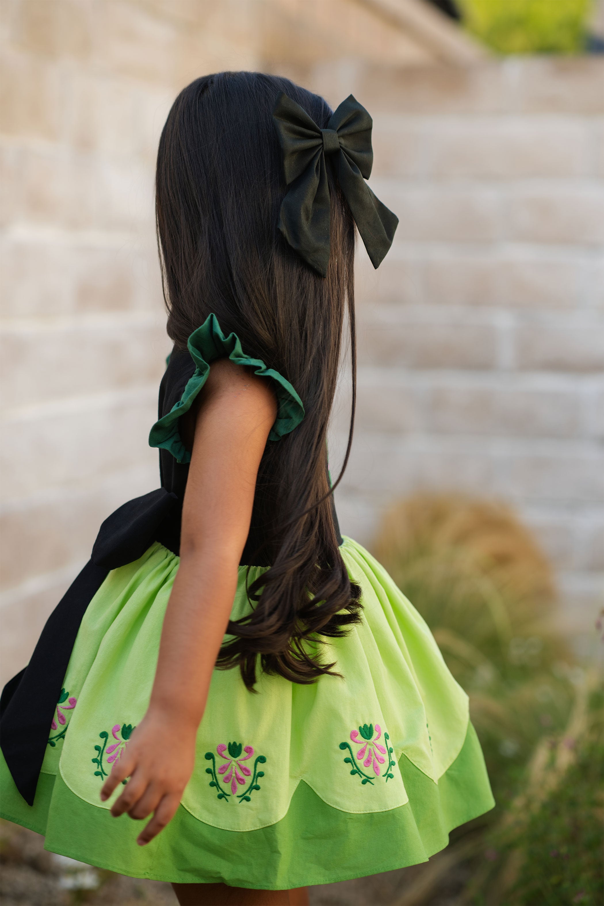 Child wearing a green dress with floral patterns and a large black bow in hair.