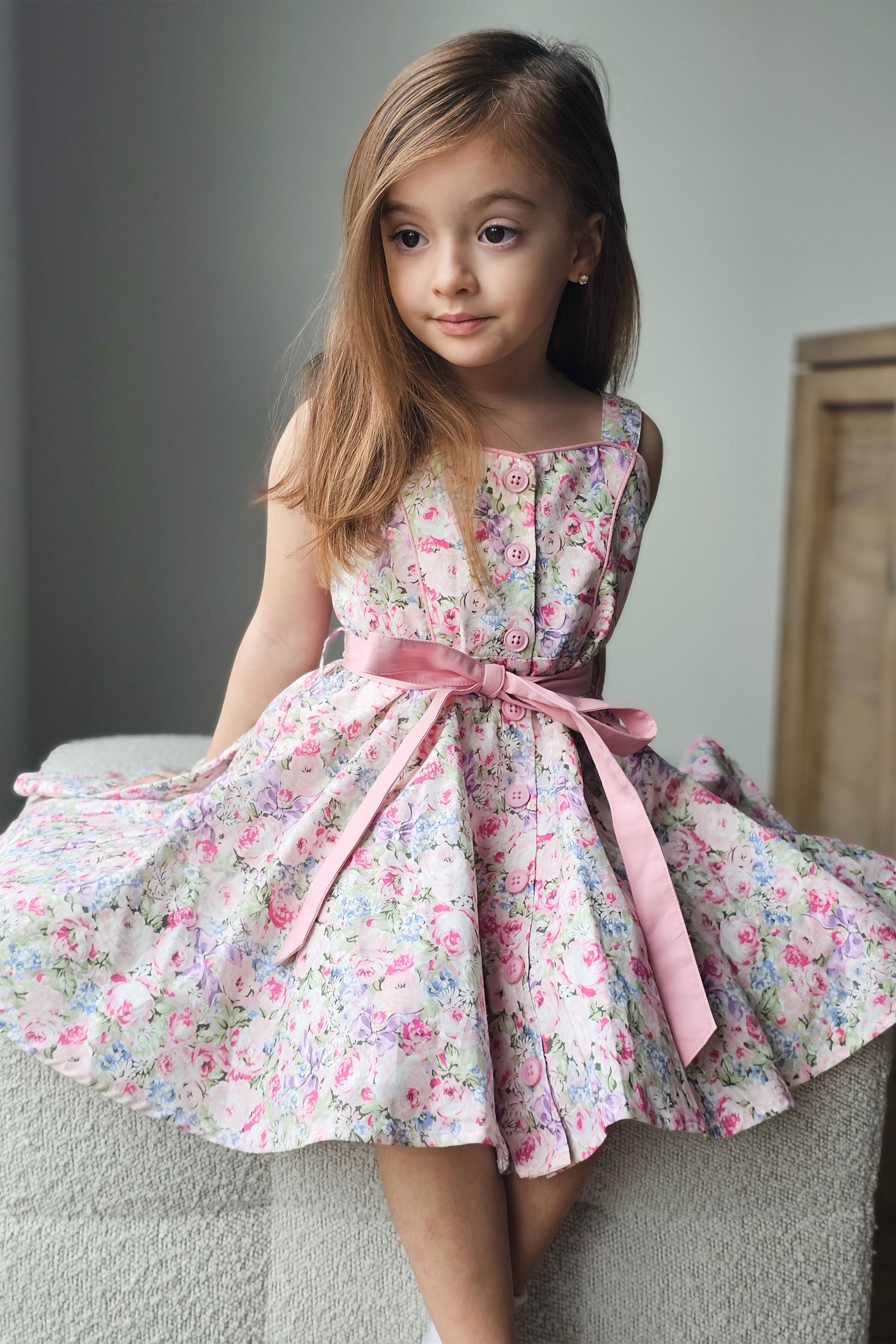 Young girl wearing a floral dress with a pink belt sitting on a couch.