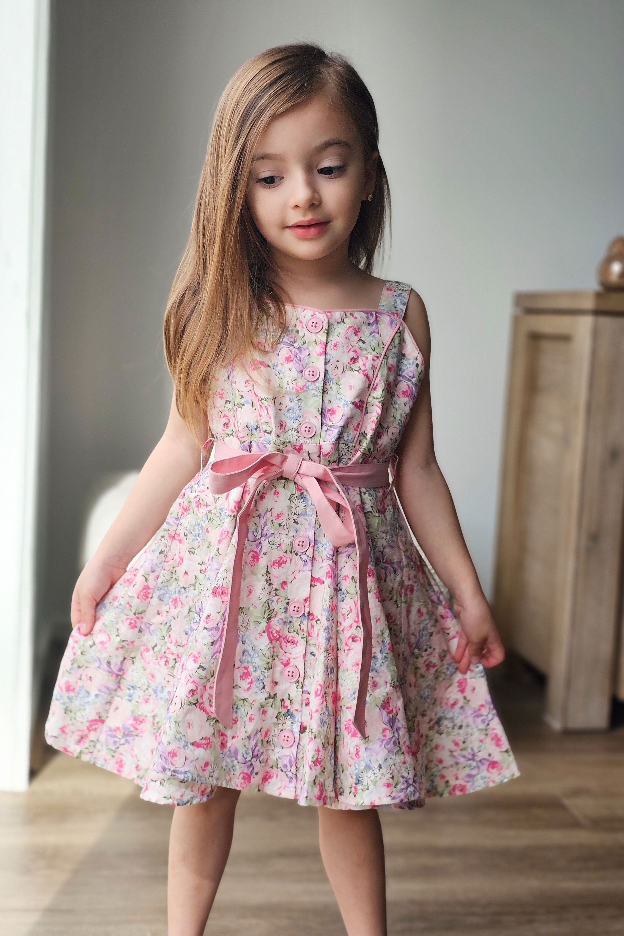 Young girl wearing a floral dress with a pink ribbon in a room.