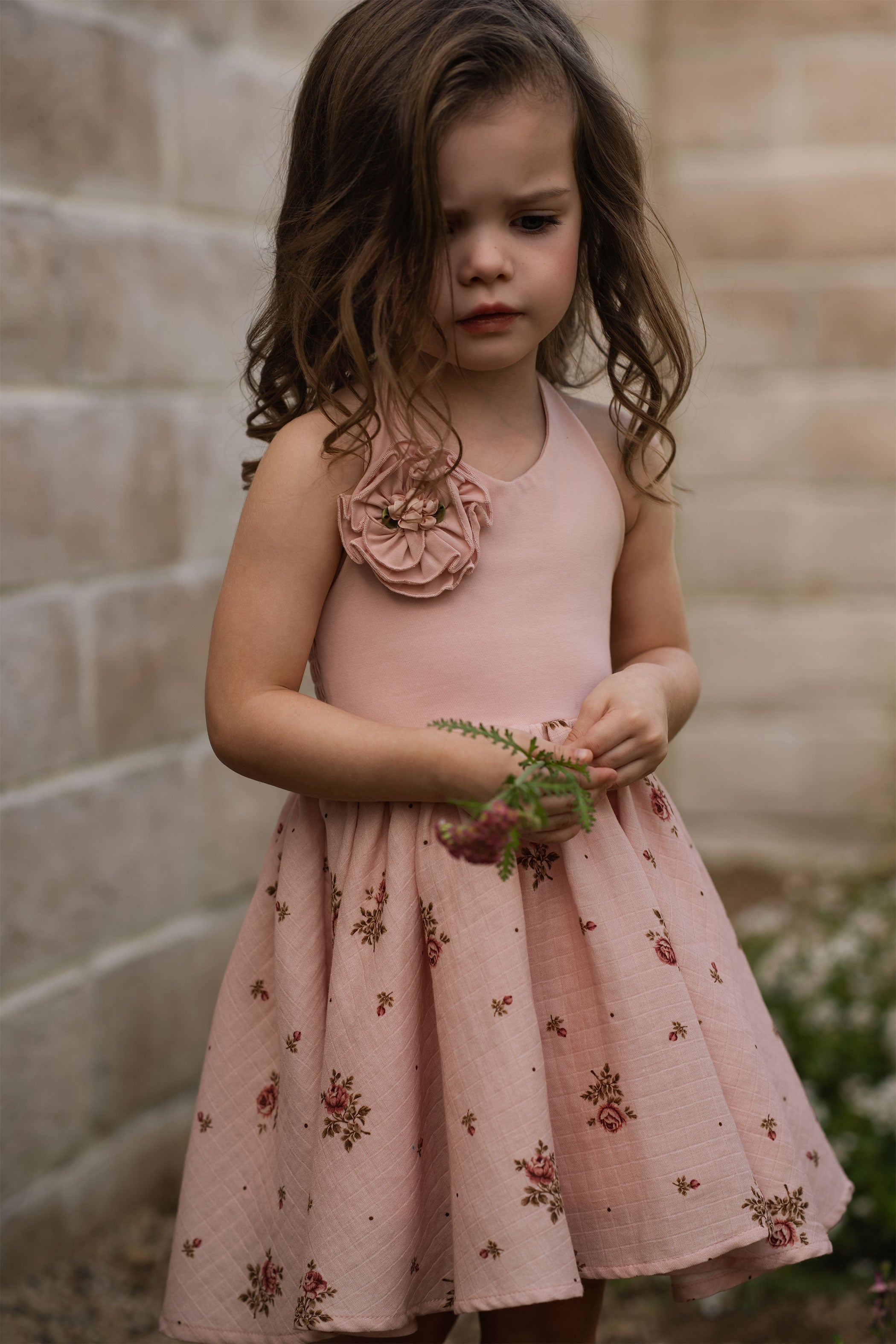 Young girl in a pink floral dress holding flowers against a stone wall.