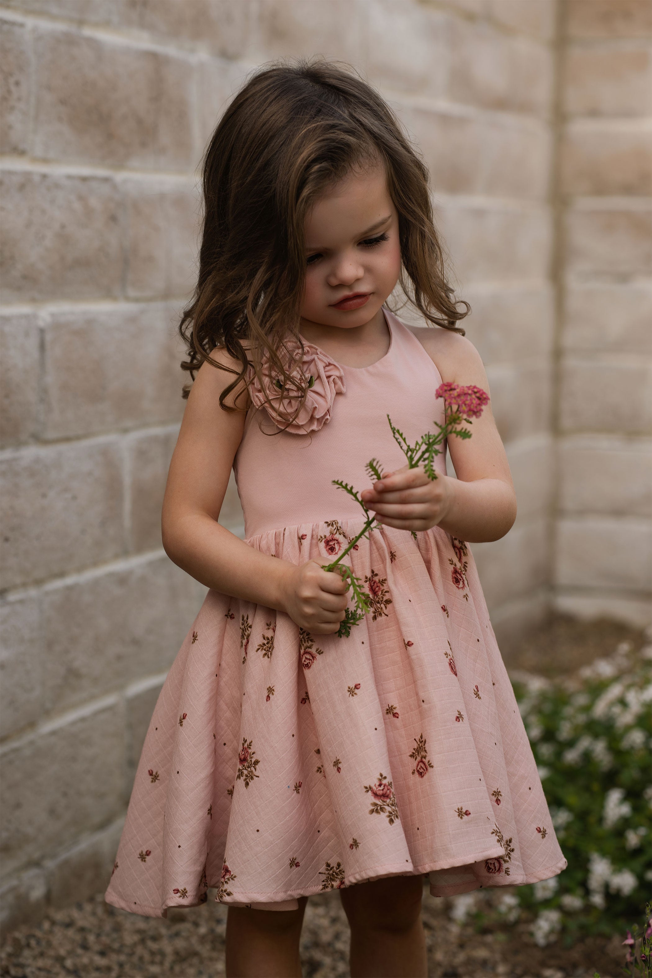 Young girl in a pink floral dress holding flowers against a stone wall.