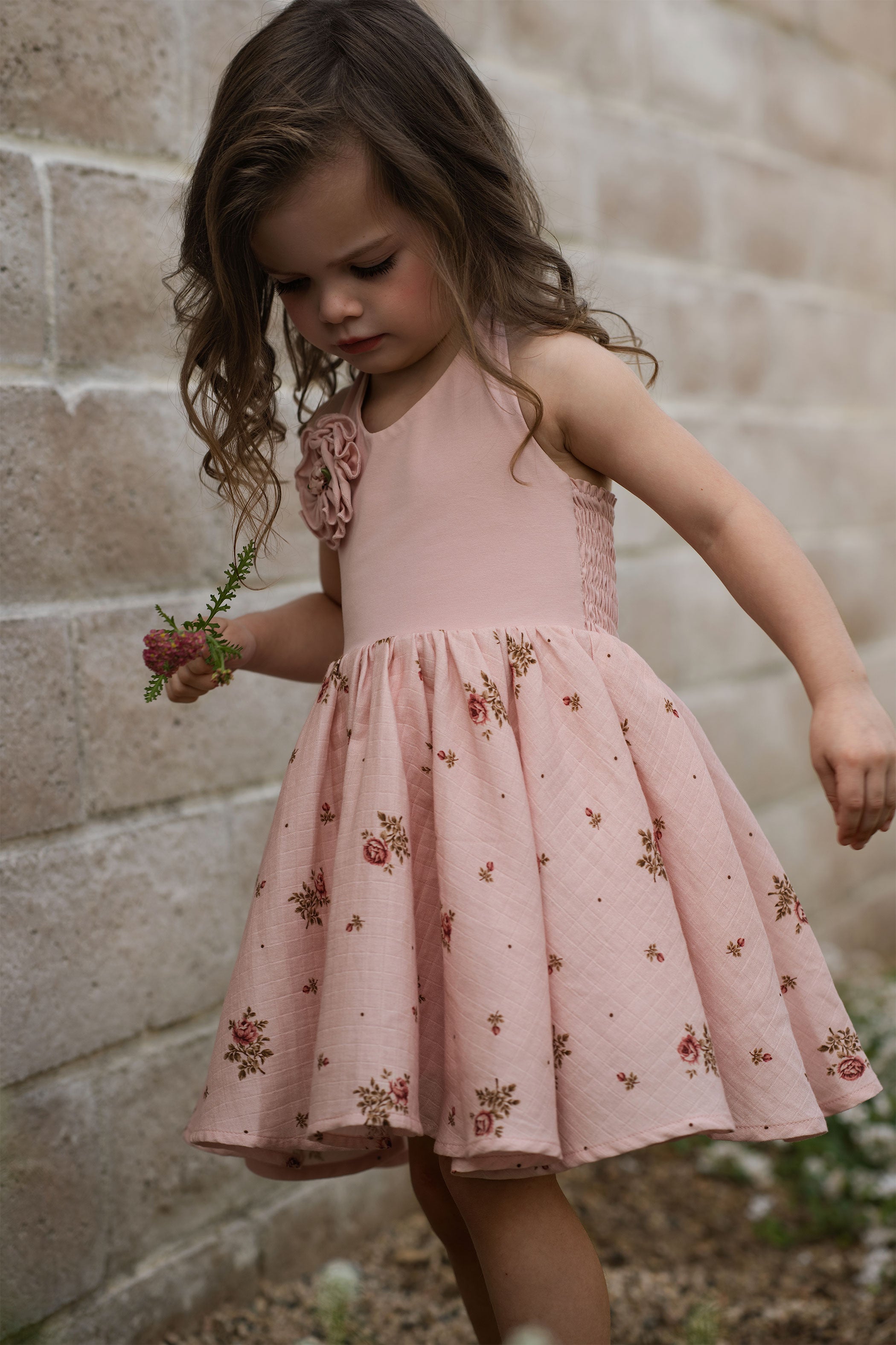Young girl in a pink floral dress standing against a stone wall.