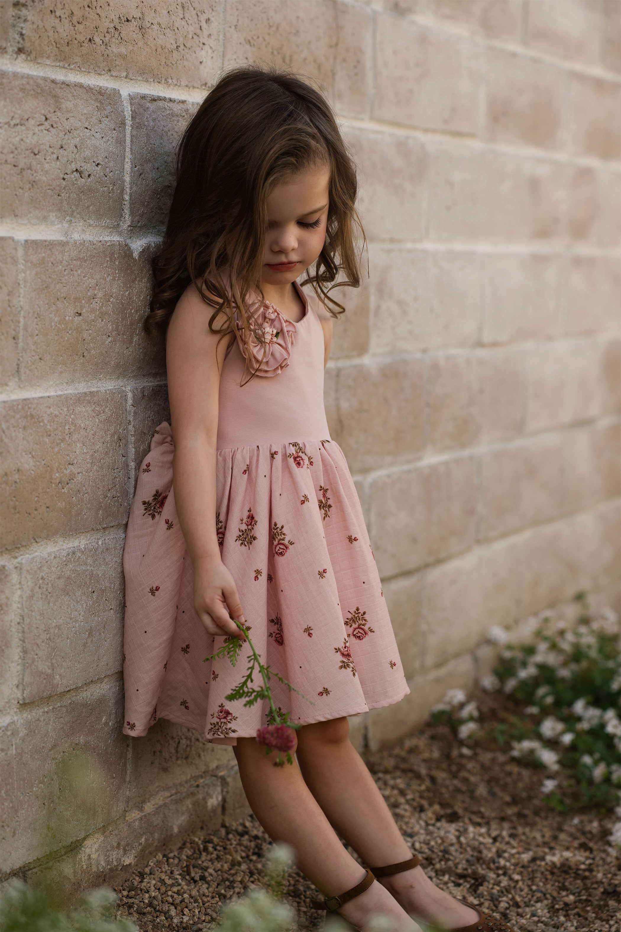 Young girl in a pink floral dress standing against a stone wall.