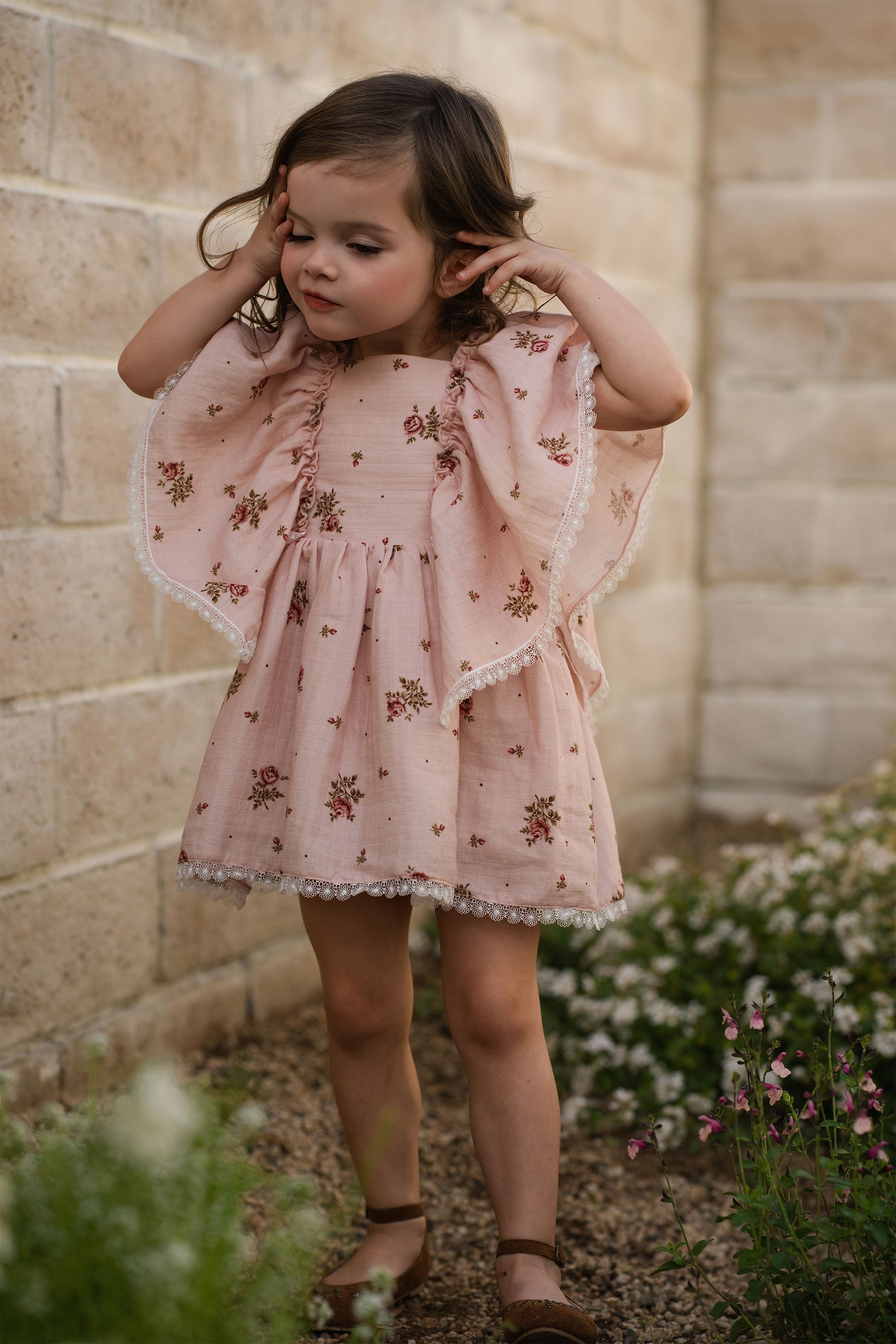 Young girl in a pink floral dress standing outdoors against a stone wall.