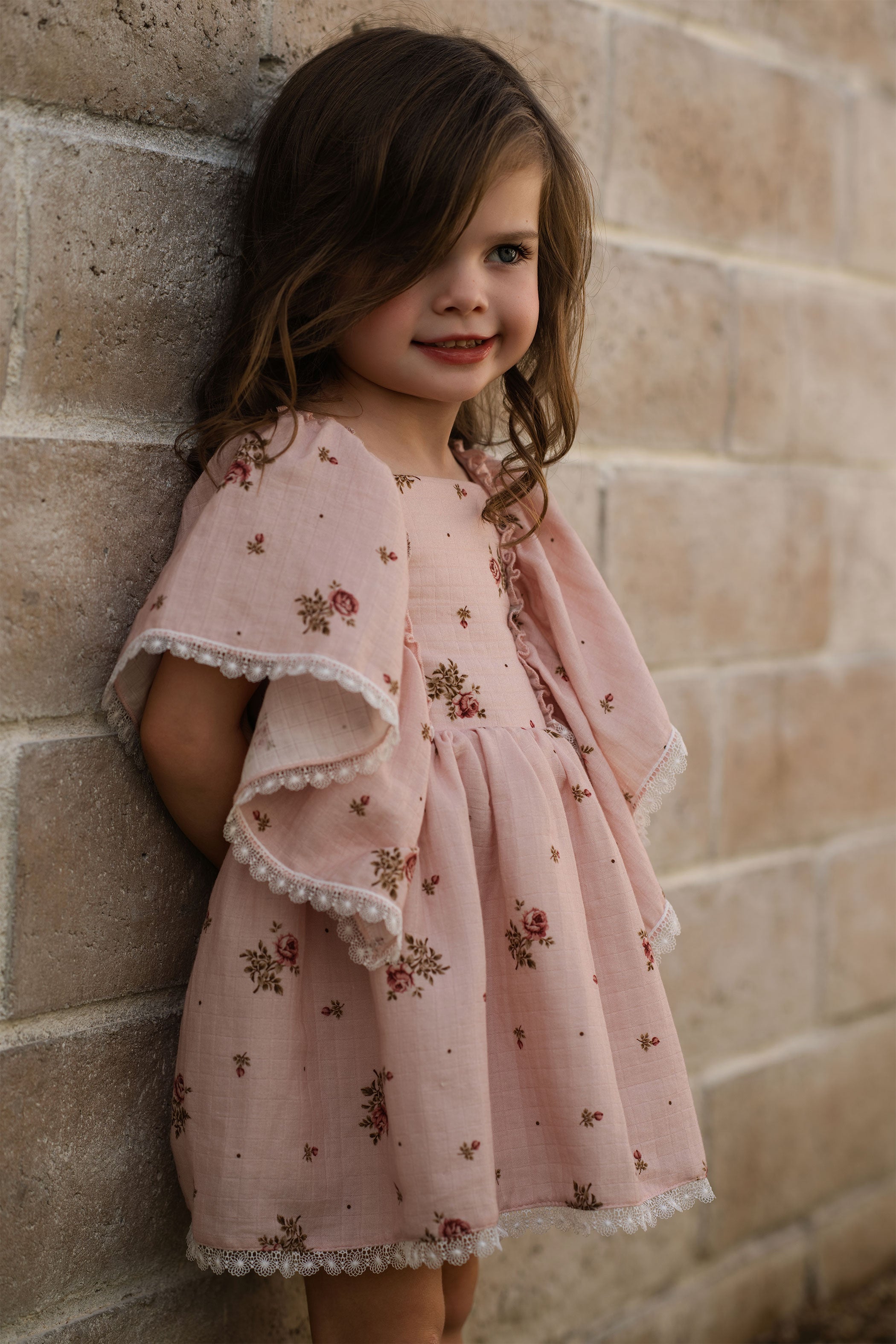 Young girl in a pink floral dress standing against a brick wall.