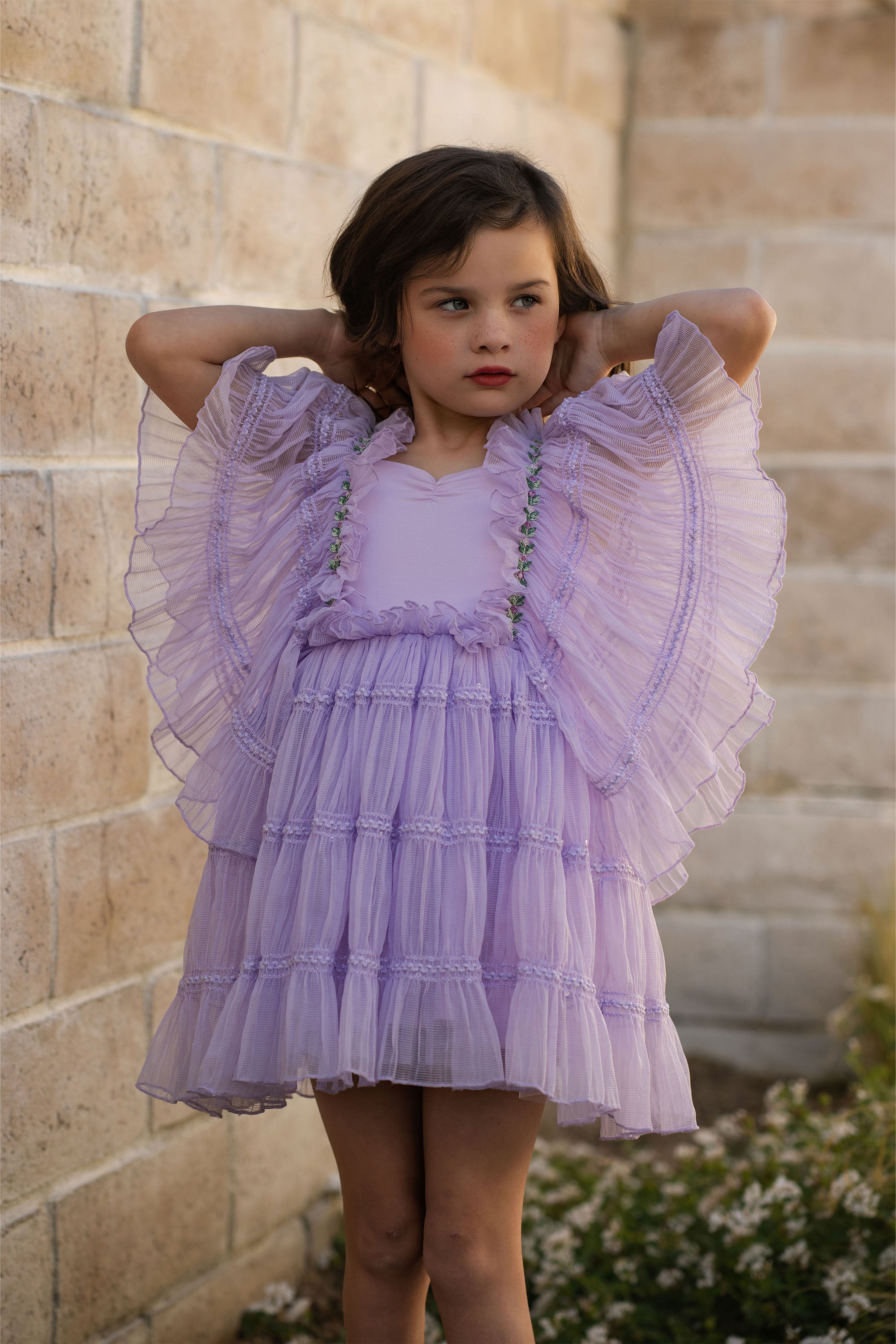 Young girl wearing a purple dress with ruffled sleeves against a stone wall.