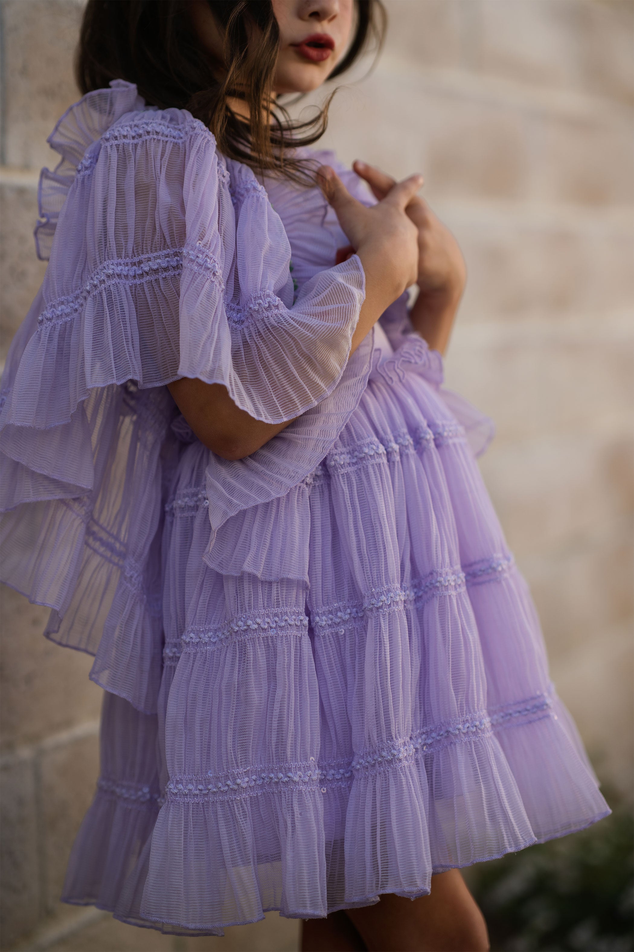 Girl wearing a light purple dress with ruffled sleeves and layers, standing against a stone wall.