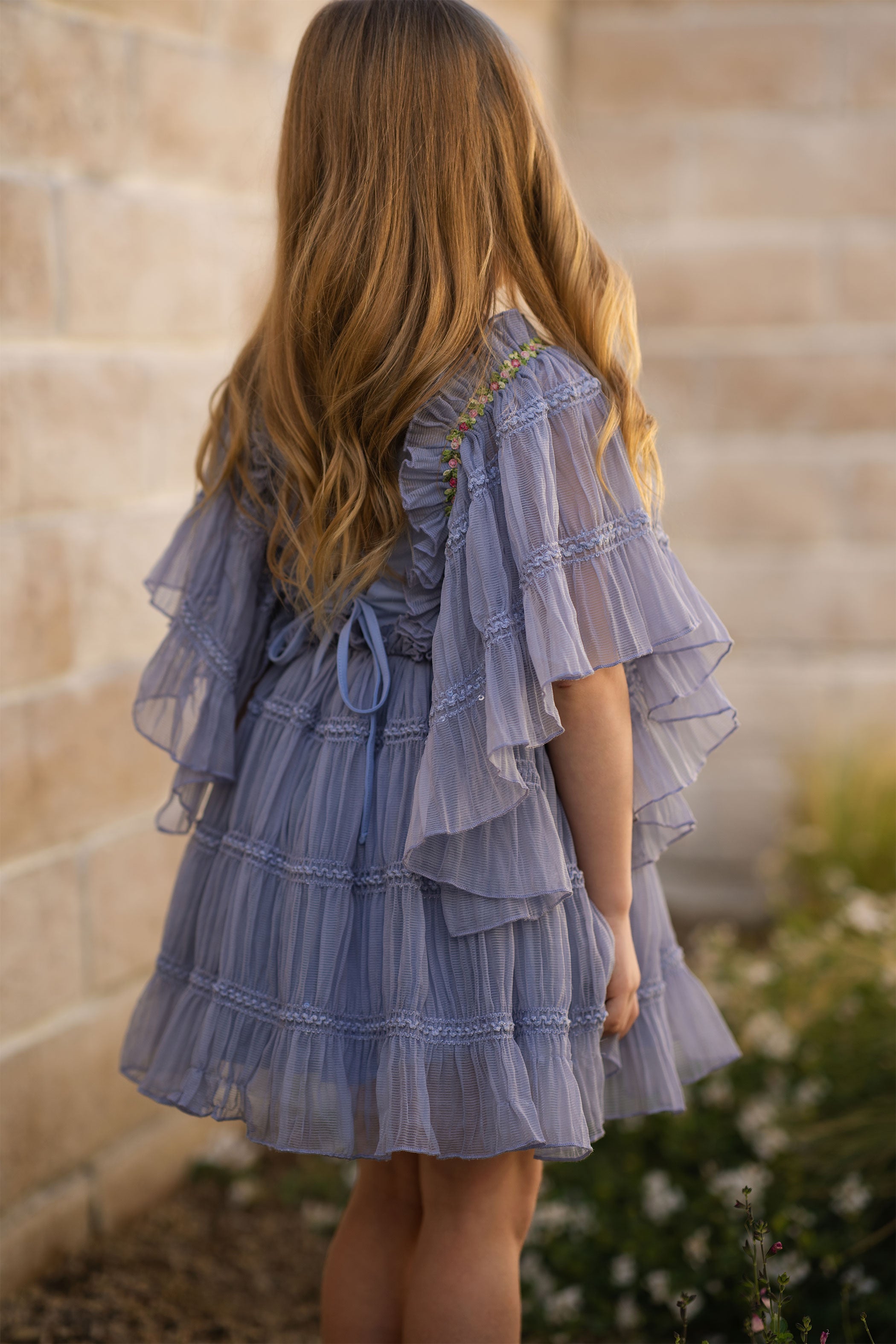 Back view of a person wearing a light blue ruffled dress against a stone wall.