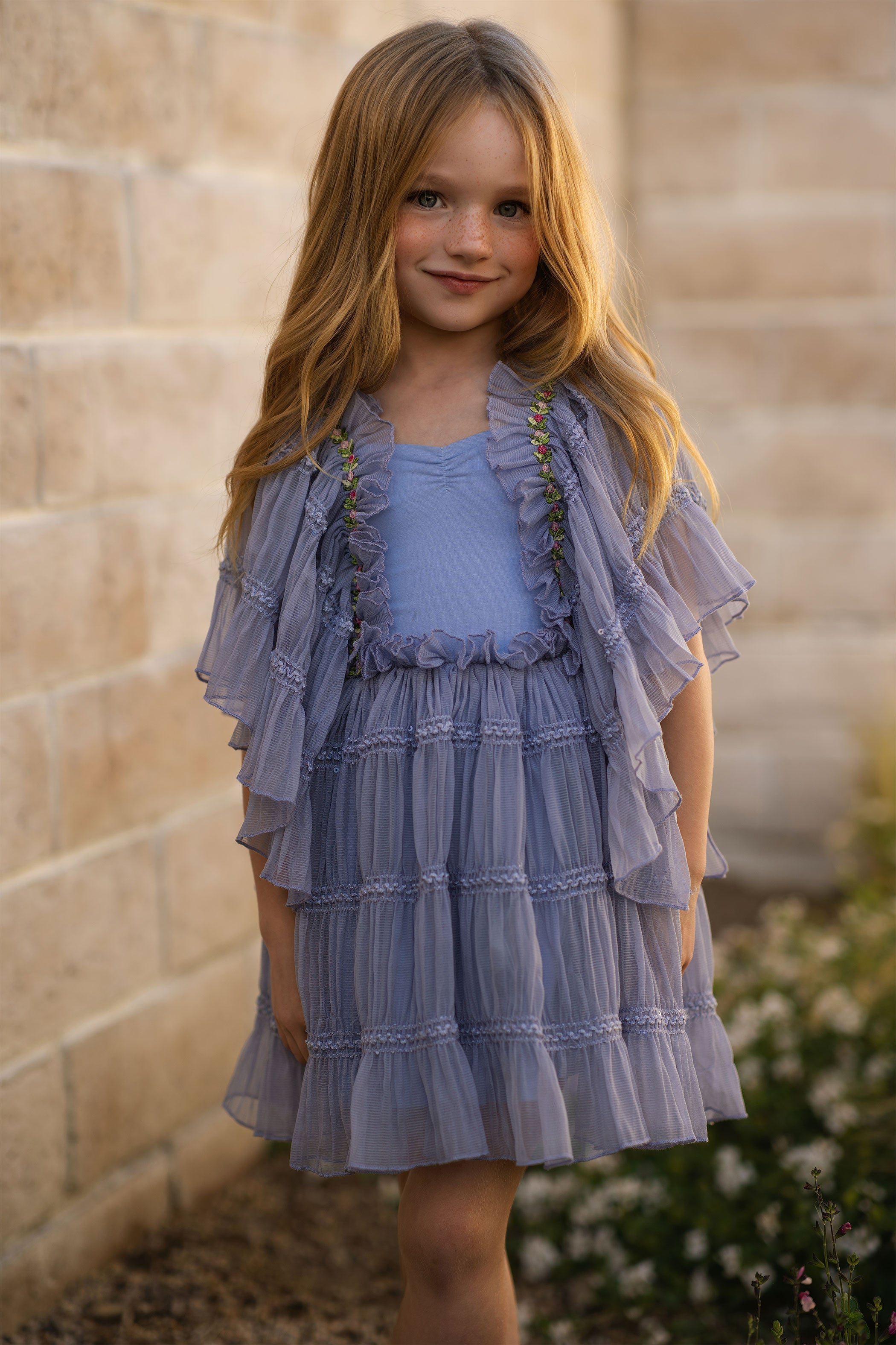 Young girl wearing a layered purple dress against a stone wall.