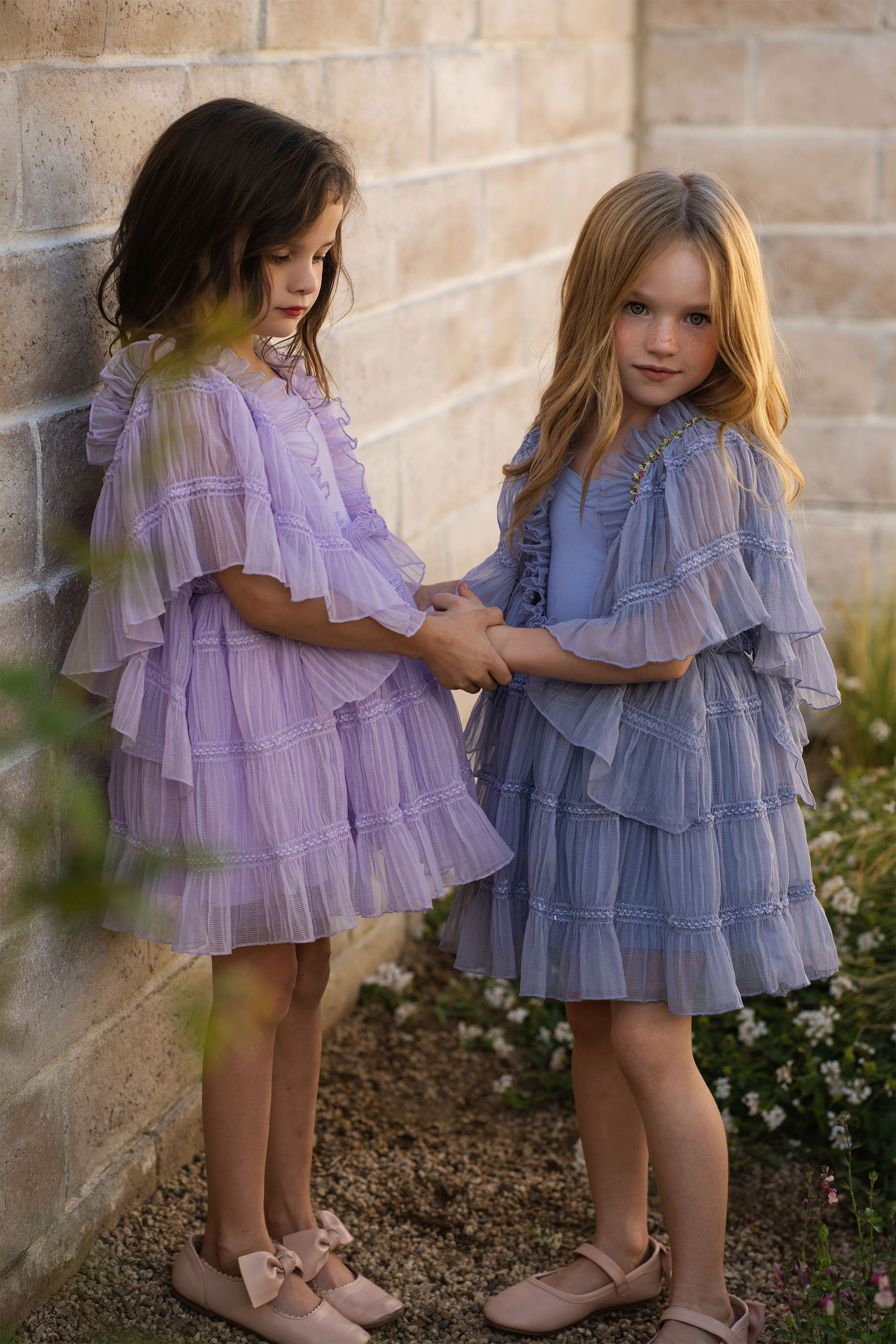 Two young girls in ruffled dresses standing against a stone wall.
