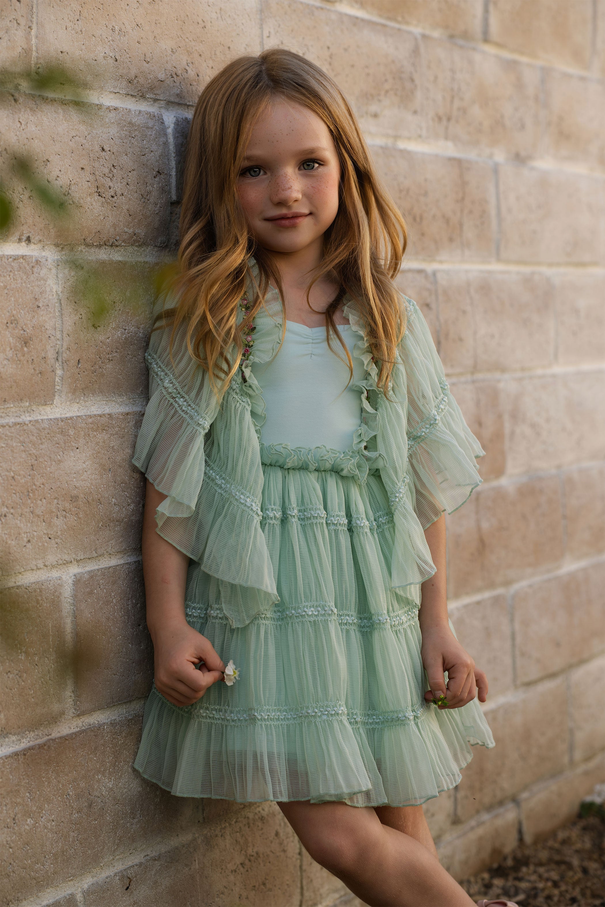 Young girl in a light green dress standing against a brick wall.
