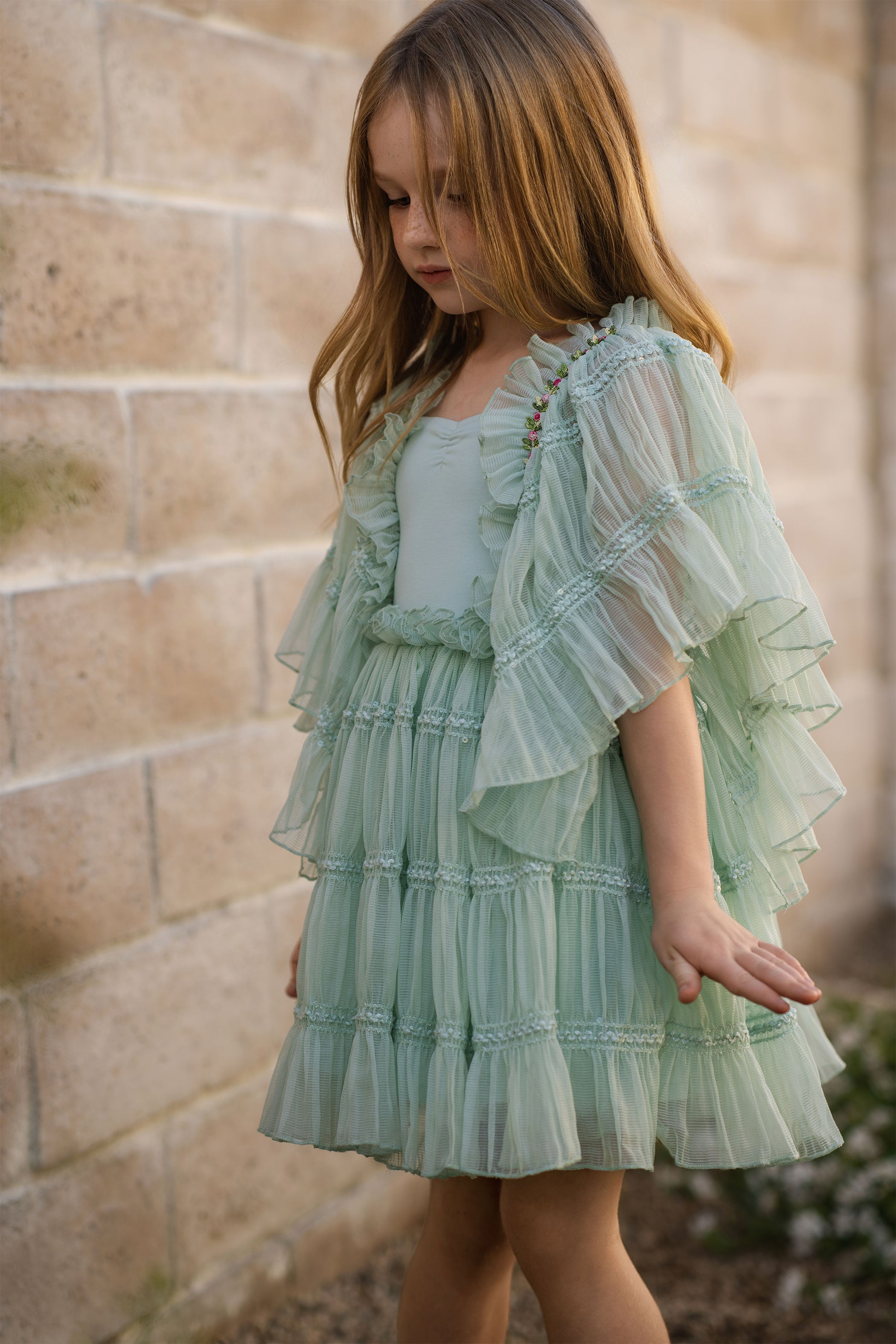 Young girl wearing a light green dress with ruffled sleeves against a stone wall.